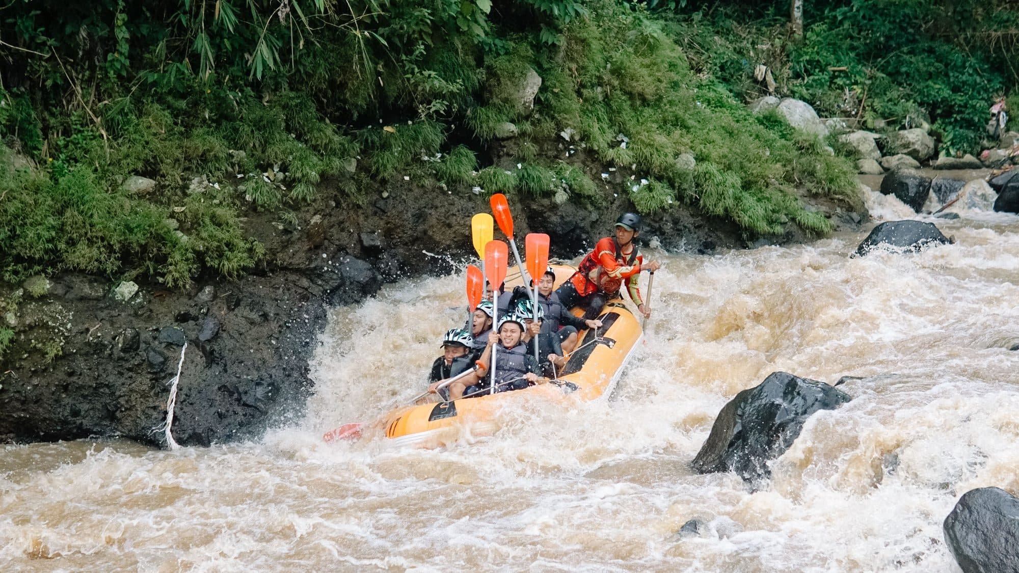 An orange raft with a guide and guests paddling through whitewater, orange paddles raised
