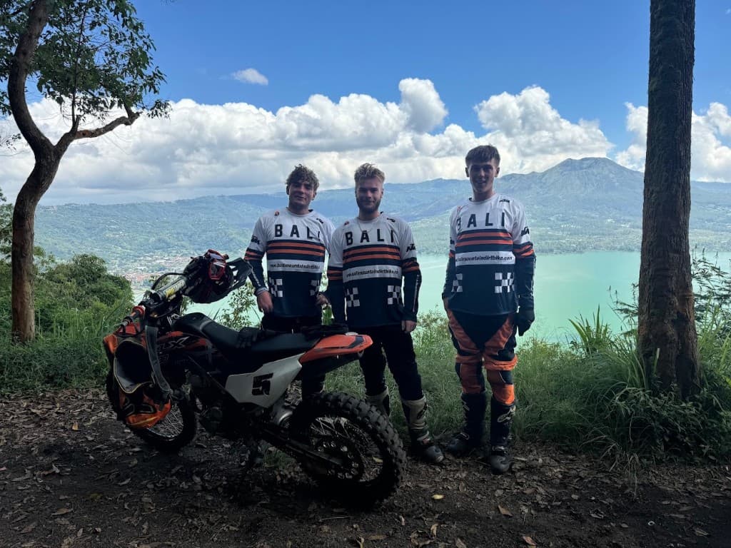 Three riders in BALI riding jerseys standing beside an orange KTM dirt bike on a ridge above Lake Batur, with Mount Batur behind