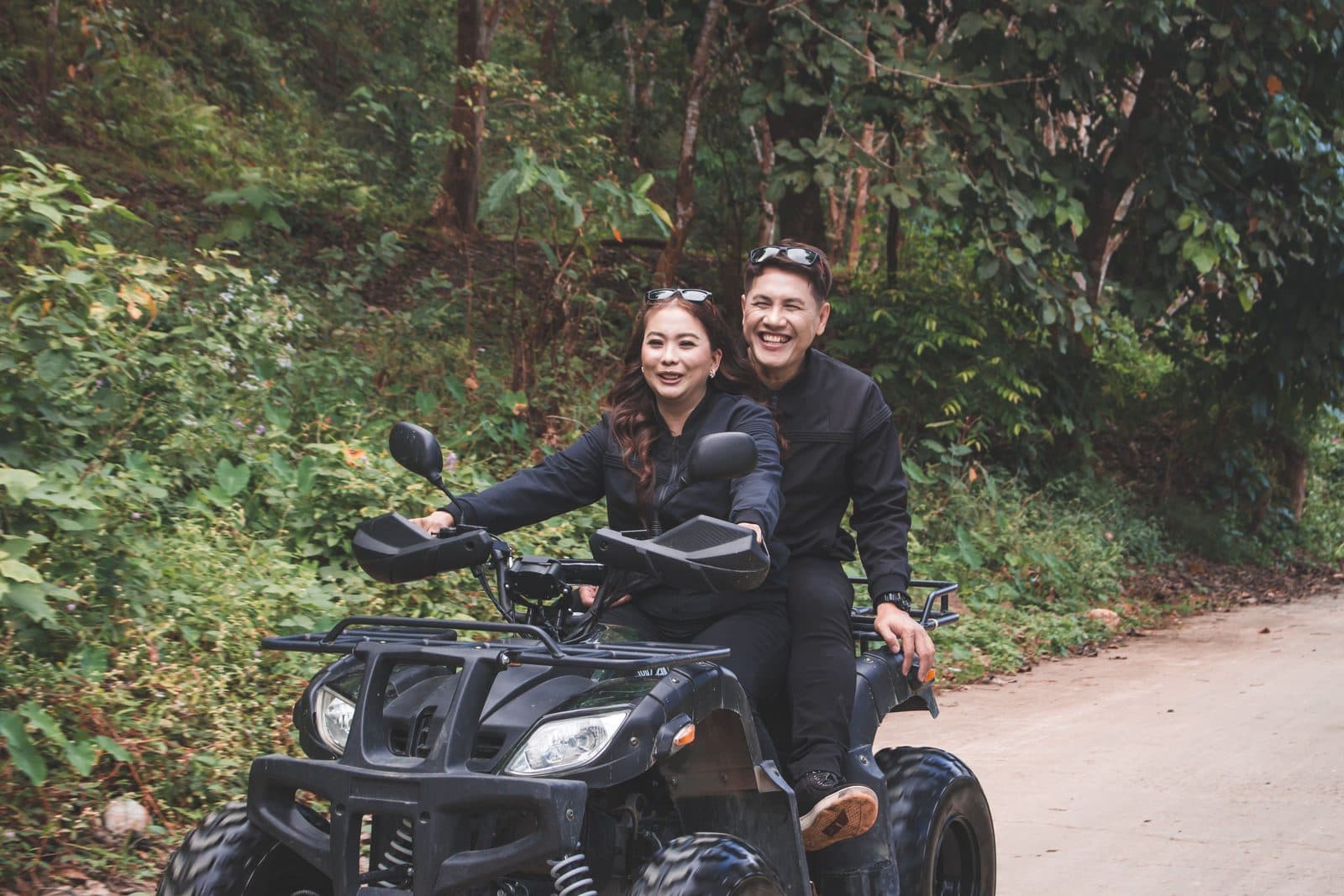 Two people in black riding one black quad on a jungle road, sunglasses on their heads